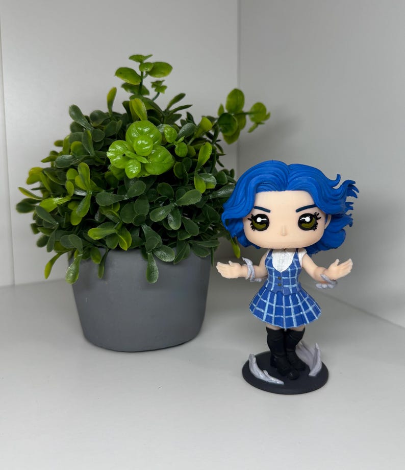 May include: A figurine of a character with blue hair and a blue and white plaid dress. The figurine has black boots and is standing on a black base with white accents. A small potted plant with green leaves is to the left.