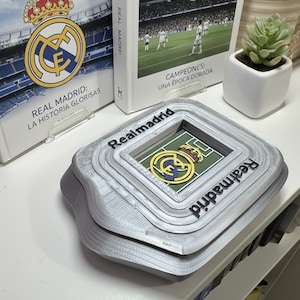 May include: A grey, layered stadium-shaped ornament with the text "Realmadrid" on the sides. The centre features a green field with a team logo. Books with the team logo and text are in the background.