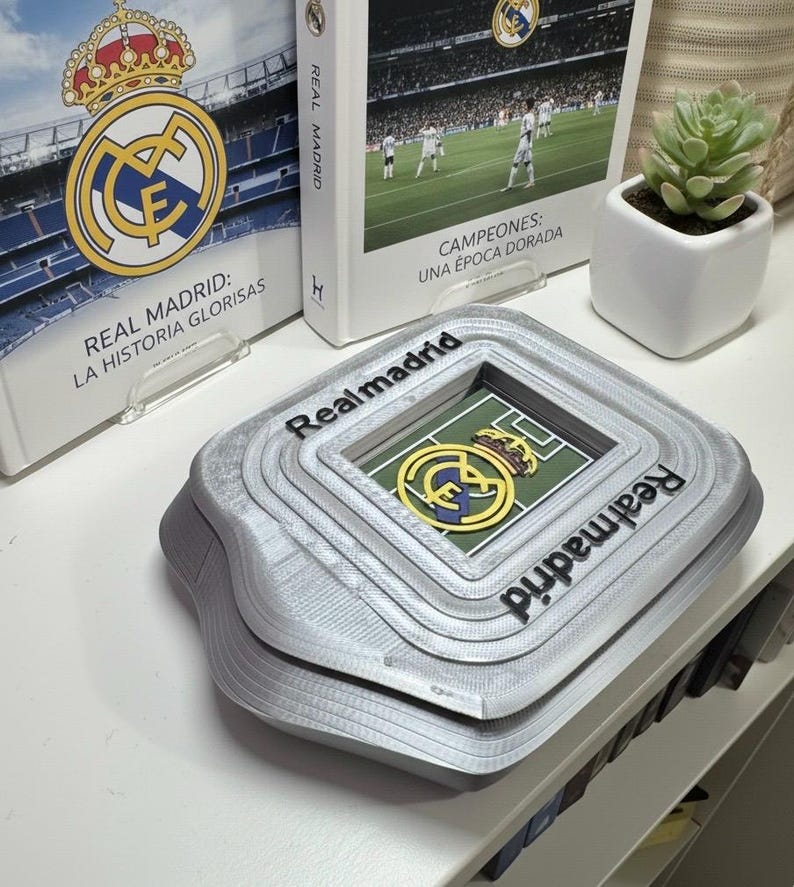 May include: A grey, layered stadium-shaped ornament with the text "Realmadrid" on the sides. The centre features a green field with a team logo. Books with the team logo and text are in the background.
