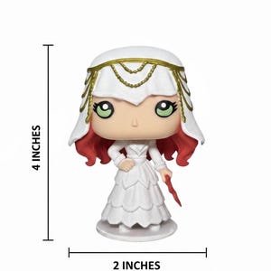 May include: A white figurine of a woman in a long dress, 4 inches tall and 2 inches wide. She has red hair, green eyes, and a white veil with gold accents. She holds a red object in her right hand. The figurine is on a white base.