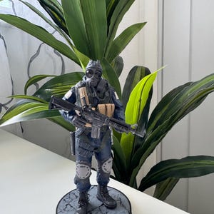May include: A detailed figurine of a soldier in a gas mask and tactical gear, holding a rifle. The figure is predominantly dark blue and black, with tan accents. It stands on a circular base with a cracked stone pattern. The background features green leafy plants.