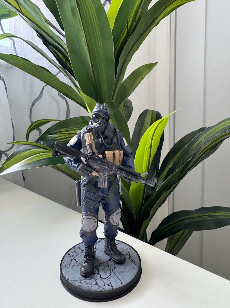 May include: A detailed figurine of a soldier in a gas mask and tactical gear, holding a rifle. The figure is predominantly dark blue and black, with tan accents. It stands on a circular base with a cracked stone pattern. The background features green leafy plants.