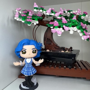 May include: A blue-haired figurine in a blue plaid dress stands before a bonsai tree constructed from building blocks. The tree features a brown trunk, green leaves, and pink blossoms. The bonsai sits on a dark brown wooden base.