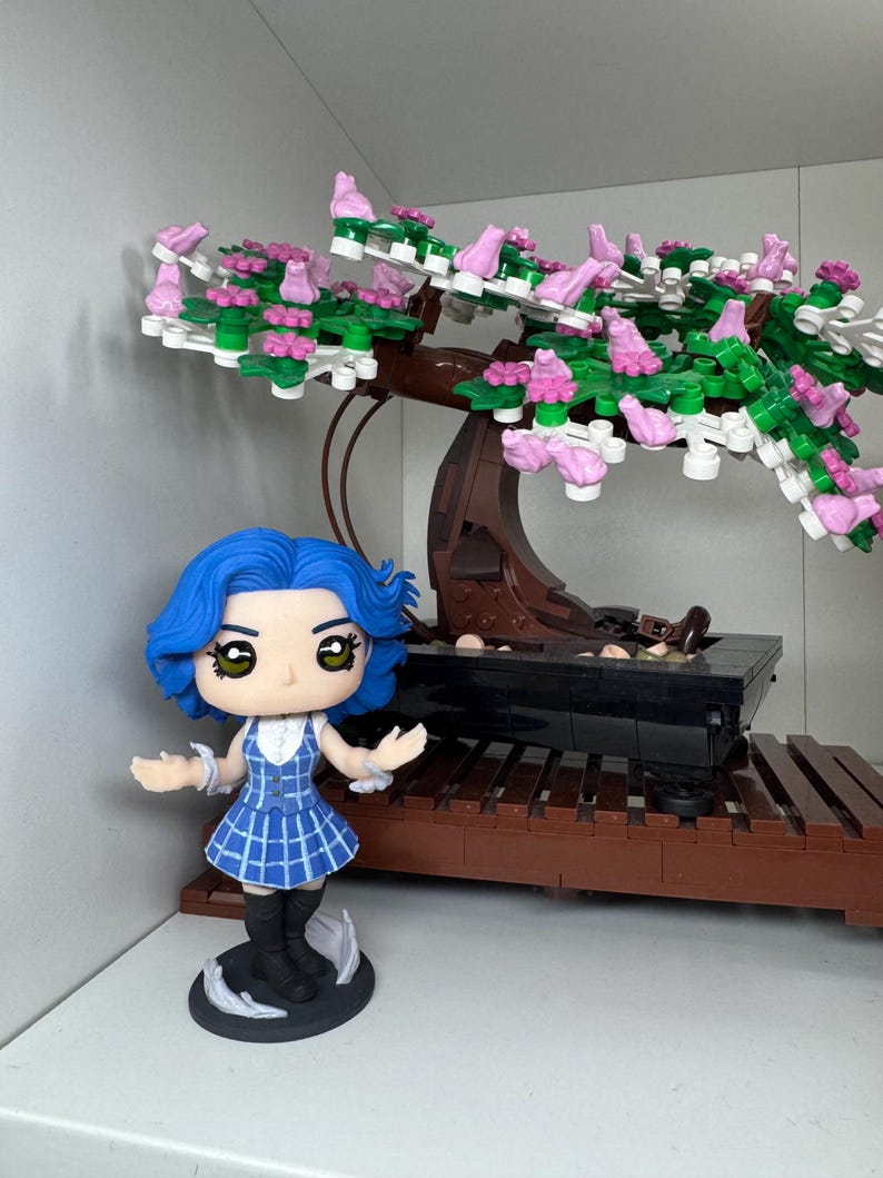 May include: A blue-haired figurine in a blue plaid dress stands before a bonsai tree constructed from building blocks. The tree features a brown trunk, green leaves, and pink blossoms. The bonsai sits on a dark brown wooden base.