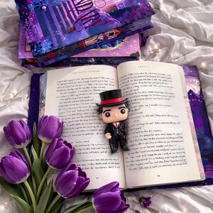 Puede incluir: Un libro abierto con texto, una figura Funko Pop con sombrero de copa y un ramo de tulipanes morados. Varios libros apilados con la palabra "Legend" en la portada son visibles. La escena est&aacute; ambientada sobre una superficie blanca.