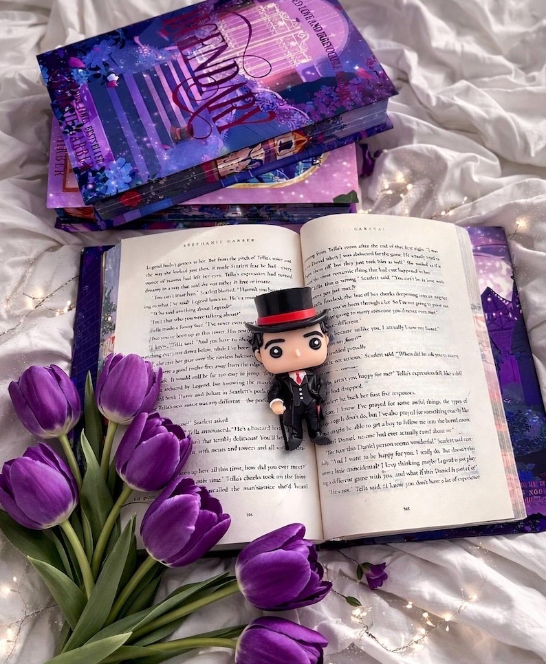 Puede incluir: Un libro abierto con texto, una figura Funko Pop con sombrero de copa y un ramo de tulipanes morados. Varios libros apilados con la palabra "Legend" en la portada son visibles. La escena est&aacute; ambientada sobre una superficie blanca.