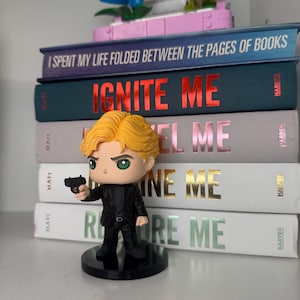 Pu&ograve; includere: Una statuina di un uomo in abito nero con una pistola, davanti a una pila di libri. I libri hanno titoli come "IGNITE ME" e "RESTORE ME". Fiori decorativi Lego sono sopra i libri.