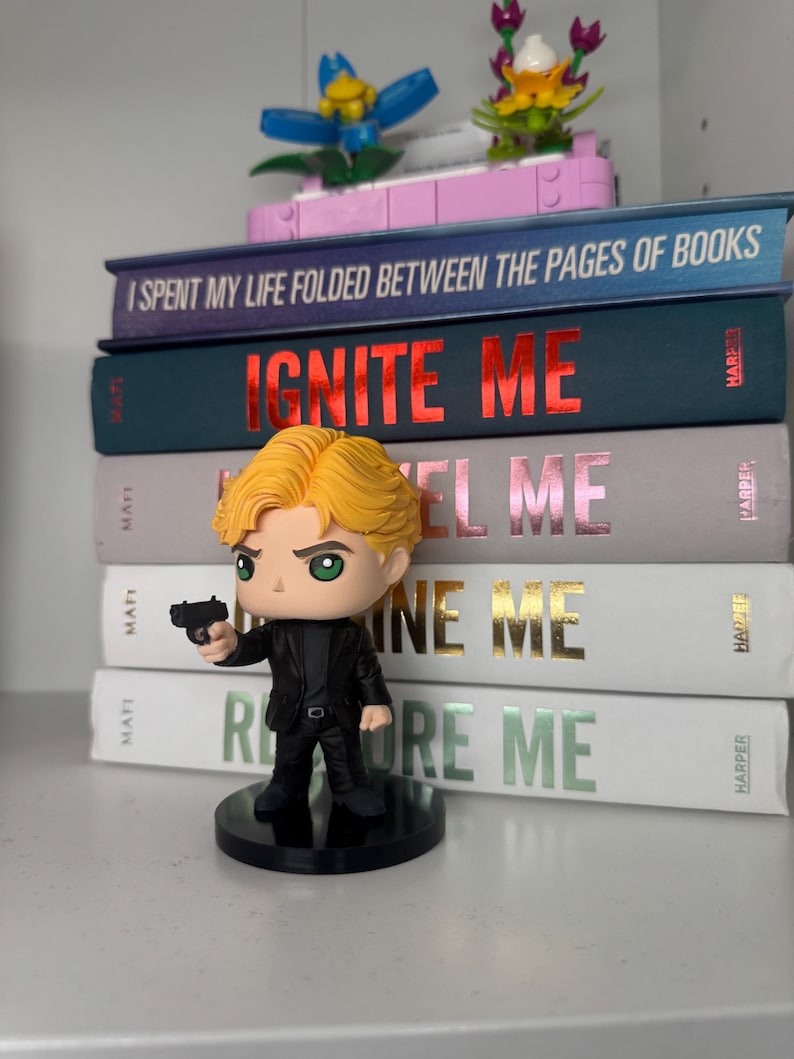 Pu&ograve; includere: Una statuina di un uomo in abito nero con una pistola, davanti a una pila di libri. I libri hanno titoli come "IGNITE ME" e "RESTORE ME". Fiori decorativi Lego sono sopra i libri.