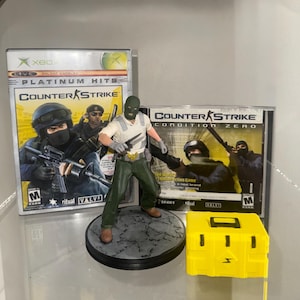 May include: An assortment of Counter-Strike themed items. Includes an Xbox game case, a PC game case, a figurine of a character holding knives, and a yellow box with a lightning bolt symbol.