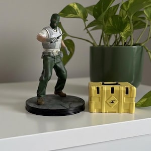 May include: A figurine of a character in a green balaclava and green pants, holding a knife, stands on a round base. A yellow box with a black handle and a diamond-shaped logo is next to it. A green potted plant is in the background.