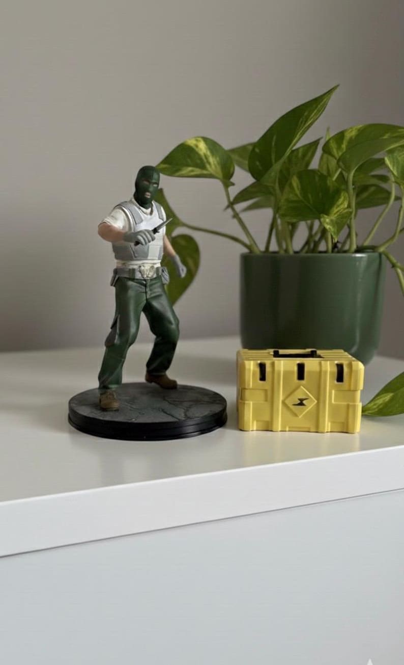 May include: A figurine of a character in a green balaclava and green pants, holding a knife, stands on a round base. A yellow box with a black handle and a diamond-shaped logo is next to it. A green potted plant is in the background.