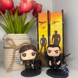 May include: A shelf display featuring two Funko Pop figures of characters from a fantasy series, with a vase of red tulips and two books. The figures are black and gold, with small dragon companions. The books have orange and yellow covers.