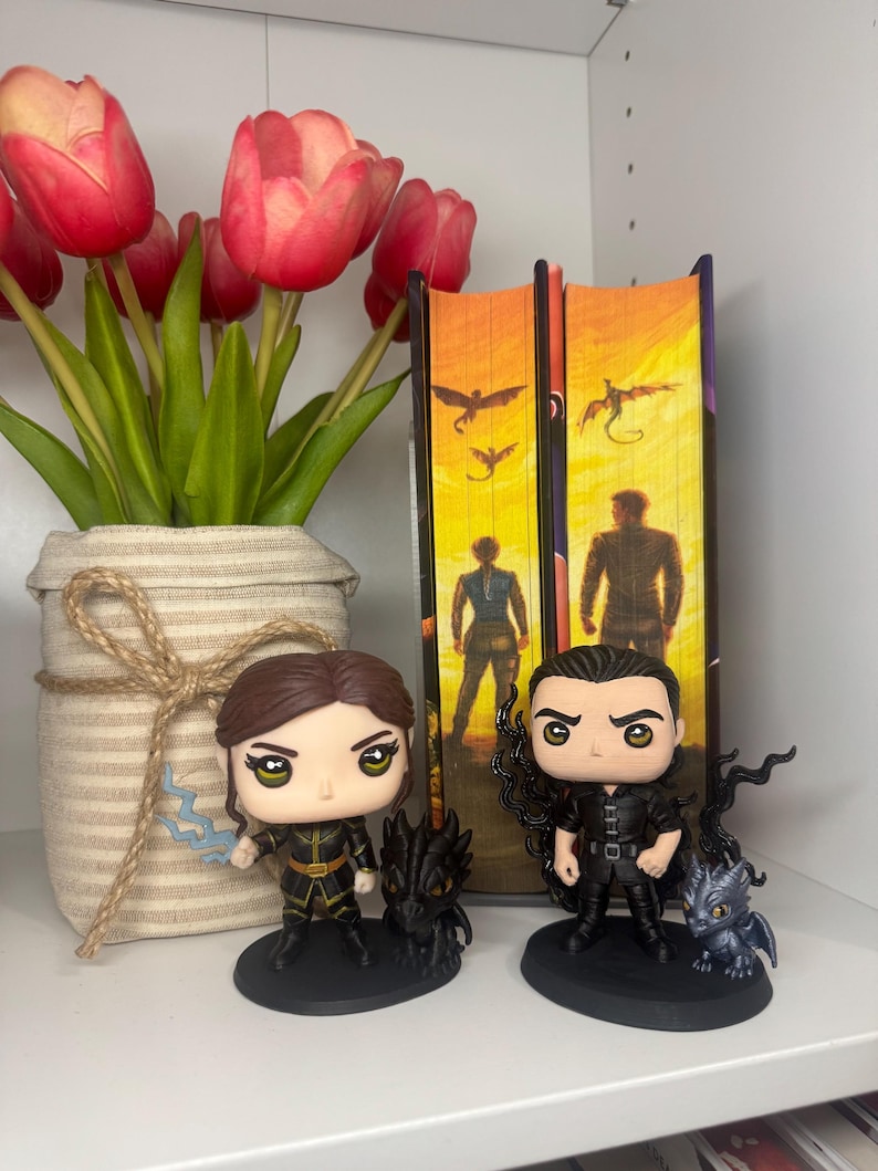 May include: A shelf display featuring two Funko Pop figures of characters from a fantasy series, with a vase of red tulips and two books. The figures are black and gold, with small dragon companions. The books have orange and yellow covers.