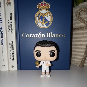 May include: A Funko Pop figure of a footballer in a white kit, holding a golden trophy. It stands before a blue book with the Real Madrid logo and the words "Coraz&oacute;n Blanco". Two books are on the left.