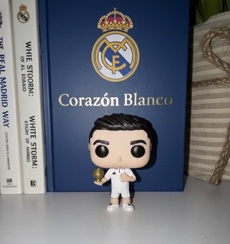 May include: A Funko Pop figure of a footballer in a white kit, holding a golden trophy. It stands before a blue book with the Real Madrid logo and the words "Coraz&oacute;n Blanco". Two books are on the left.