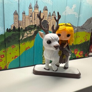K&ouml;nnte beinhalten: Eine wei&szlig;e Figur eines Hirsches mit gro&szlig;en Geweihen, die eine blondhaarige Figur mit Krone und braunem Outfit tr&auml;gt. Die Figur steht auf einem braunen Sockel. Der Hintergrund zeigt ein Wandgem&auml;lde eines Schlosses und einer Landschaft.