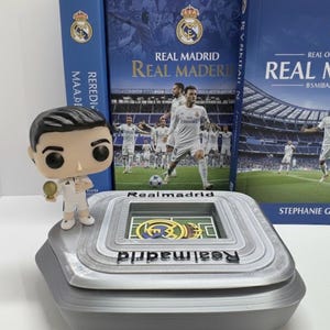 May include: A Real Madrid themed collectible set. It includes a stadium model, a figurine, and two books. The books have blue covers with the Real Madrid logo and team photos. The stadium model is grey with the Real Madrid logo.