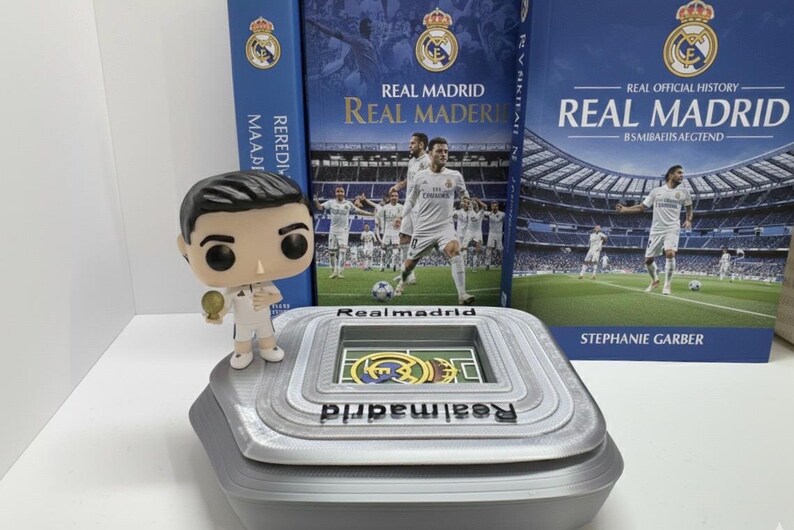 May include: A Real Madrid themed collectible set. It includes a stadium model, a figurine, and two books. The books have blue covers with the Real Madrid logo and team photos. The stadium model is grey with the Real Madrid logo.