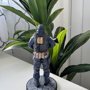 May include: A figurine of a soldier in a dark blue uniform, complete with tactical gear and a helmet. The figure is standing on a circular base with a cracked stone design. The background features green leafy plants.