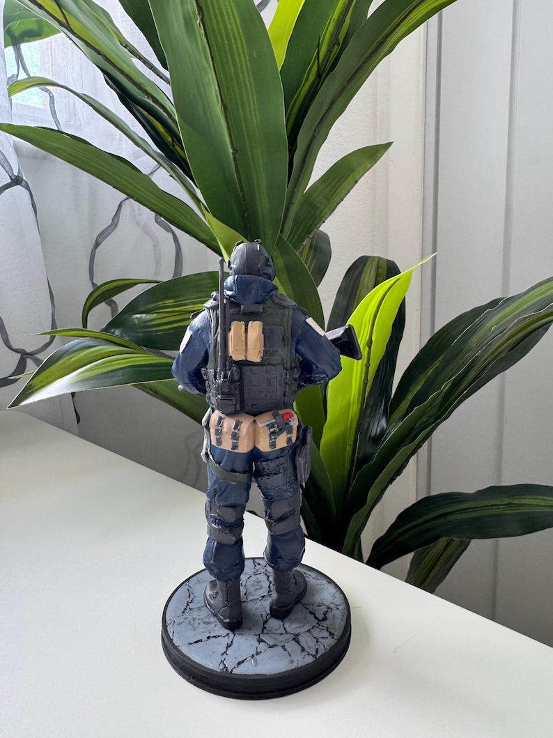 May include: A figurine of a soldier in a dark blue uniform, complete with tactical gear and a helmet. The figure is standing on a circular base with a cracked stone design. The background features green leafy plants.
