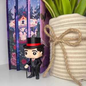 Puede incluir: Una figura Funko Pop de un hombre con traje negro, sombrero de copa y bast&oacute;n se encuentra frente a libros decorativos y un jarr&oacute;n con tulipanes. La figura tiene una corbata roja y una banda roja en el sombrero. Los libros tienen un dise&ntilde;o de carrusel.