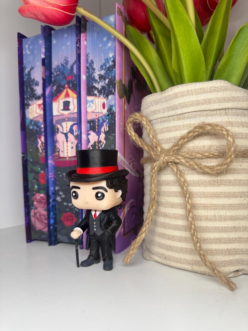 Puede incluir: Una figura Funko Pop de un hombre con traje negro, sombrero de copa y bast&oacute;n se encuentra frente a libros decorativos y un jarr&oacute;n con tulipanes. La figura tiene una corbata roja y una banda roja en el sombrero. Los libros tienen un dise&ntilde;o de carrusel.