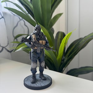 May include: A detailed figurine of a soldier in tactical gear, including a gas mask and combat boots. The figure is holding a rifle and stands on a circular base with a textured surface. The soldier's uniform is primarily dark blue with tan and gray accents.