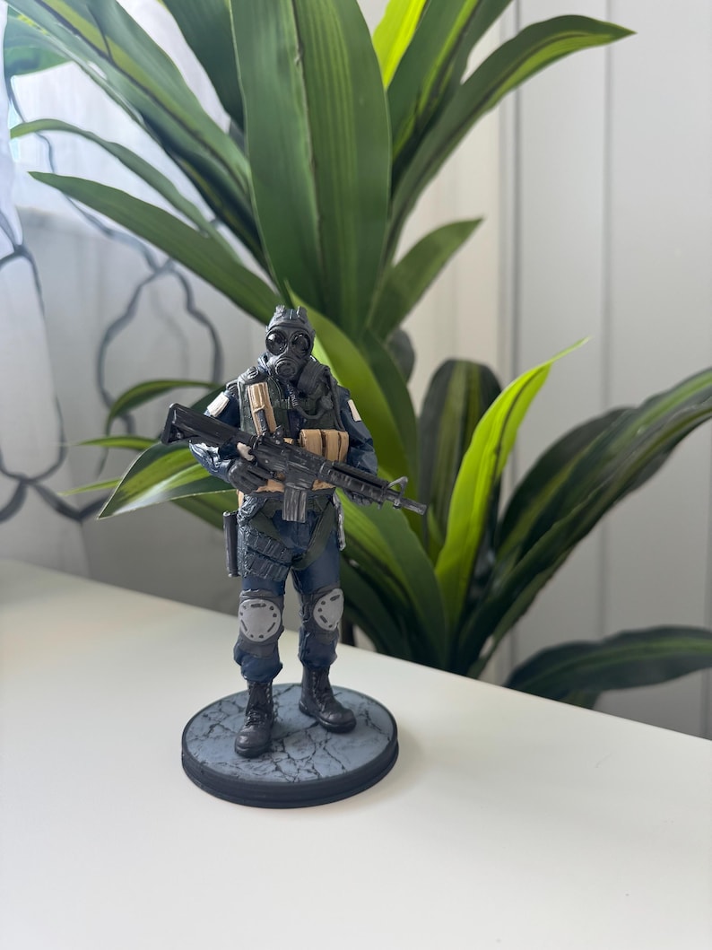 May include: A detailed figurine of a soldier in tactical gear, including a gas mask and combat boots. The figure is holding a rifle and stands on a circular base with a textured surface. The soldier's uniform is primarily dark blue with tan and gray accents.