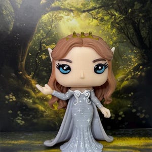 May include: A Funko Pop figure of a fantasy character with brown hair, blue eyes, and a gold crown. The figure wears a light blue dress with a cape and star details. The background features a forest scene.