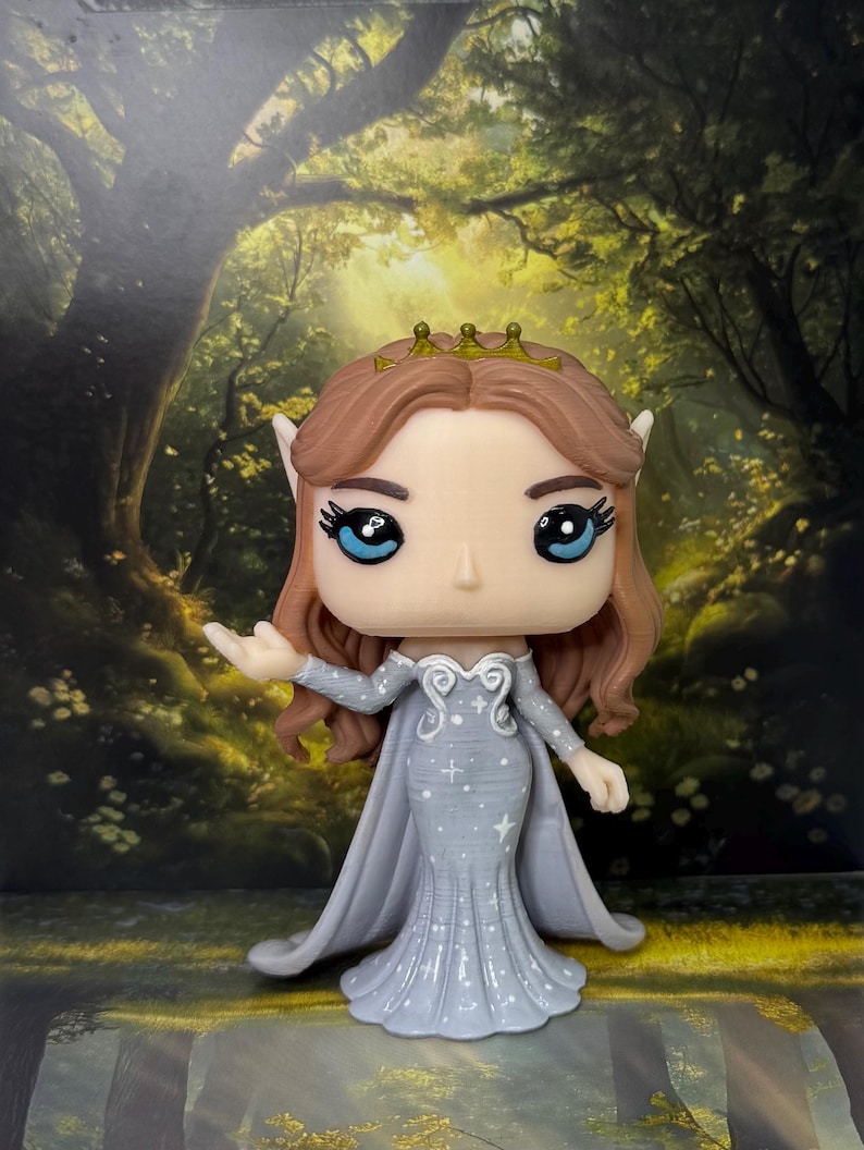 May include: A Funko Pop figure of a fantasy character with brown hair, blue eyes, and a gold crown. The figure wears a light blue dress with a cape and star details. The background features a forest scene.