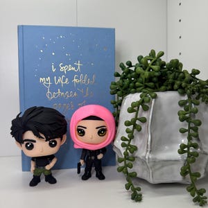 Pu&ograve; includere: Due statuette in vinile di personaggi con grandi occhi, uno con i capelli neri e l'altro con un copricapo rosa, sono davanti a un libro blu con testo dorato che recita "i spent my life folded between the pages of". Un vaso di ceramica bianco con piante verdi &egrave; a destra.