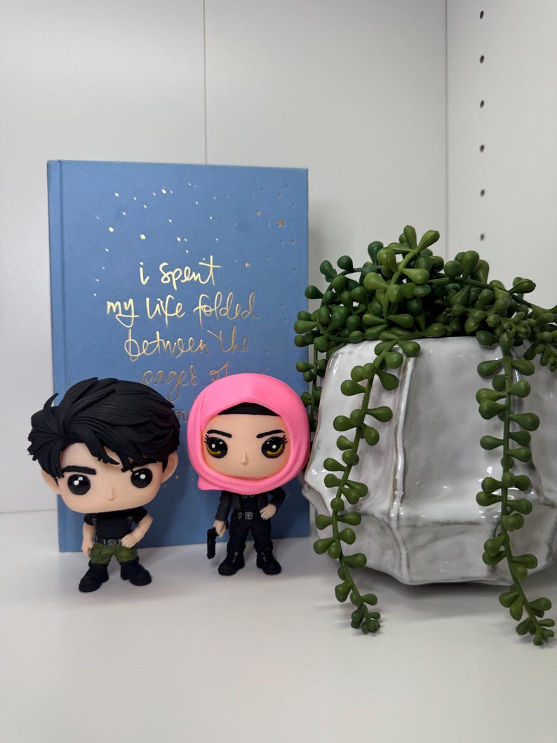 Pu&ograve; includere: Due statuette in vinile di personaggi con grandi occhi, uno con i capelli neri e l'altro con un copricapo rosa, sono davanti a un libro blu con testo dorato che recita "i spent my life folded between the pages of". Un vaso di ceramica bianco con piante verdi &egrave; a destra.