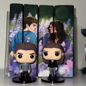 Pu&ograve; includere: Due statuette Funko Pop di personaggi di una serie di libri sono in piedi davanti alle copertine dei libri. Le statuette sono su basi circolari nere. Le copertine dei libri presentano illustrazioni di un uomo e una donna in un campo di fiori viola.