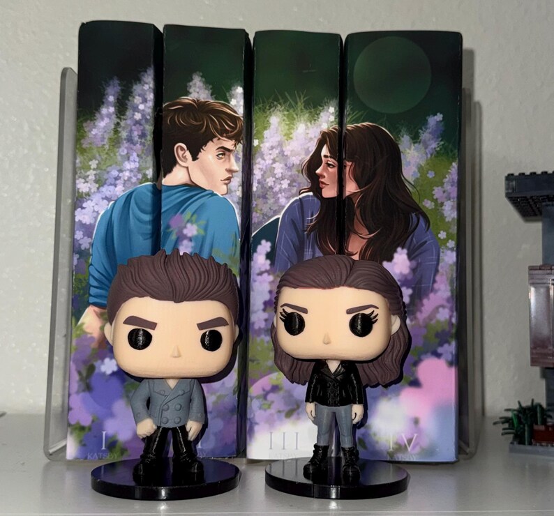 Pu&ograve; includere: Due statuette Funko Pop di personaggi di una serie di libri sono in piedi davanti alle copertine dei libri. Le statuette sono su basi circolari nere. Le copertine dei libri presentano illustrazioni di un uomo e una donna in un campo di fiori viola.