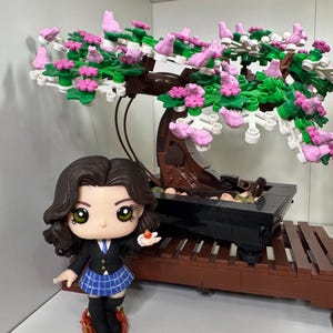 May include: A miniature figure in a school uniform, holding a small red object, stands beside a Lego bonsai tree with pink and green foliage. The bonsai is in a black pot on a brown Lego stand.