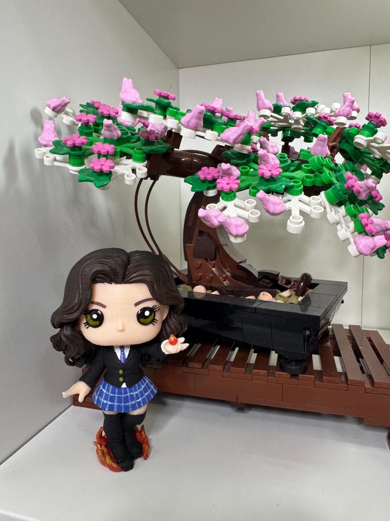 May include: A miniature figure in a school uniform, holding a small red object, stands beside a Lego bonsai tree with pink and green foliage. The bonsai is in a black pot on a brown Lego stand.