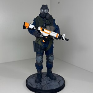 May include: A figurine of a soldier in a gas mask and tactical gear, holding a rifle with white, orange, and black accents. The figure is on a circular base with a cracked stone design. The soldier is wearing a blue uniform.