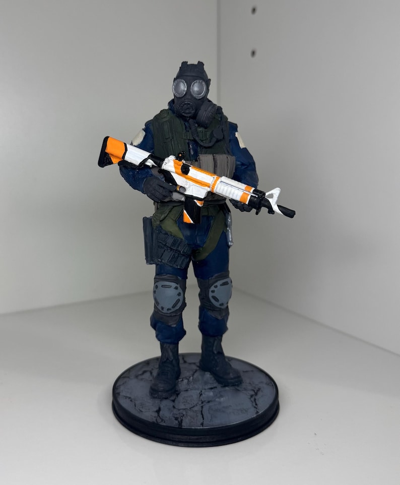 May include: A figurine of a soldier in a gas mask and tactical gear, holding a rifle with white, orange, and black accents. The figure is on a circular base with a cracked stone design. The soldier is wearing a blue uniform.