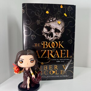 May include: A book titled "The Book Azrael" by Amber V. Nicole, with a skull and floral design on the cover, stands next to a figurine of a woman in a burgundy dress holding a flame. The figurine is on a black base.