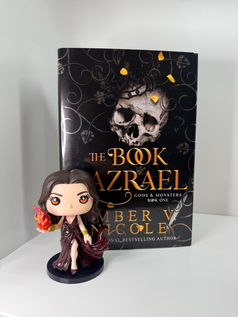 May include: A book titled "The Book Azrael" by Amber V. Nicole, with a skull and floral design on the cover, stands next to a figurine of a woman in a burgundy dress holding a flame. The figurine is on a black base.