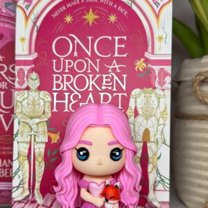 K&ouml;nnte beinhalten: Eine rosa Figur eines M&auml;dchens mit rosa Haaren und einem Kleid, das einen kleinen orangefarbenen Fuchs h&auml;lt. Die Figur steht vor einem Buch mit dem Titel "Once Upon a Broken Heart". Das Buchcover ist rosa mit wei&szlig;en und goldenen Akzenten.
