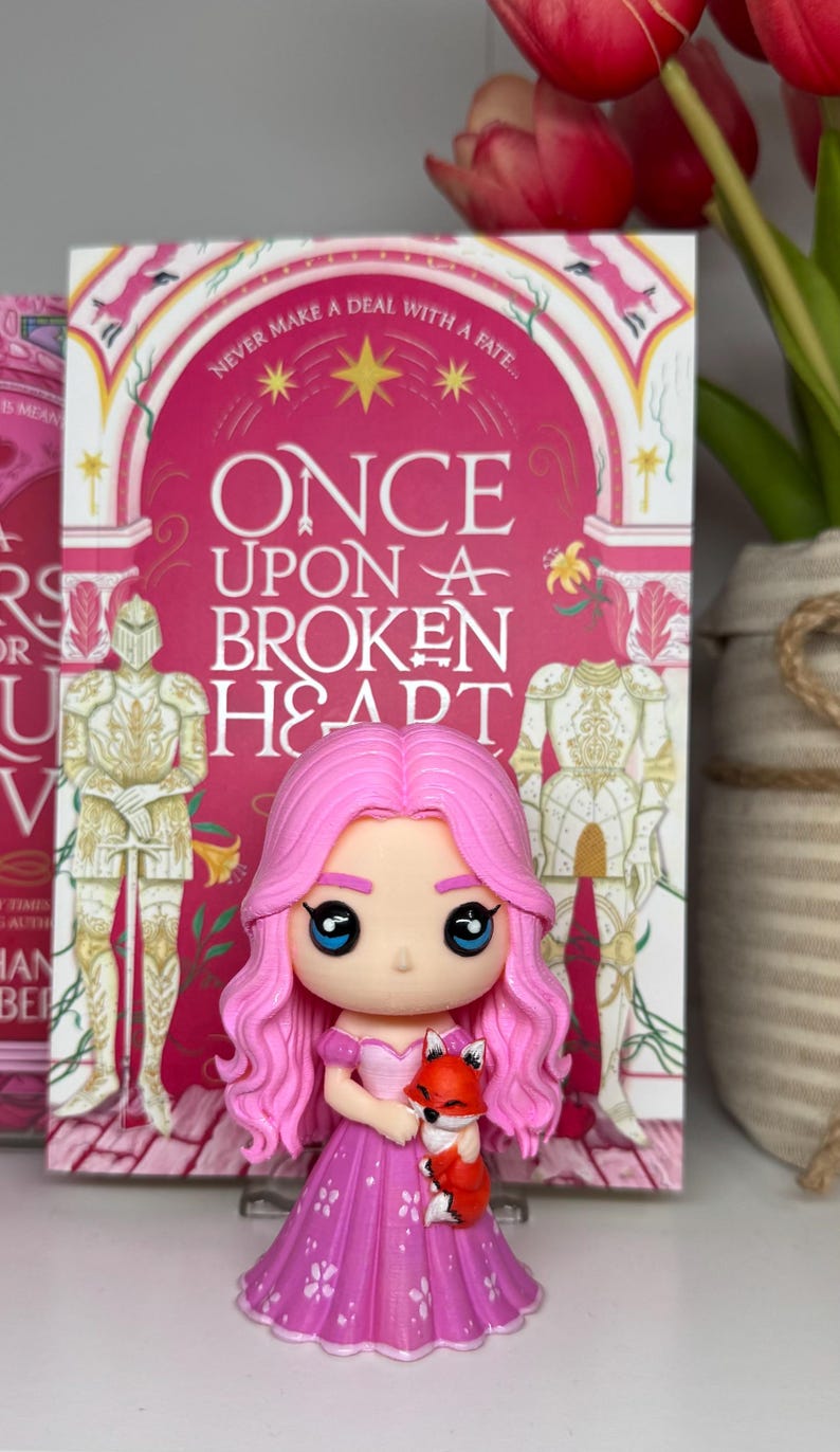 K&ouml;nnte beinhalten: Eine rosa Figur eines M&auml;dchens mit rosa Haaren und einem Kleid, das einen kleinen orangefarbenen Fuchs h&auml;lt. Die Figur steht vor einem Buch mit dem Titel "Once Upon a Broken Heart". Das Buchcover ist rosa mit wei&szlig;en und goldenen Akzenten.