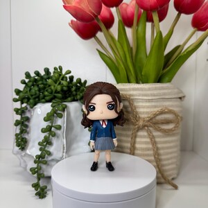 K&ouml;nnte beinhalten: Eine Figur einer Person in einem blauen Blazer, grauem Rock und schwarzen Schuhen steht auf einer wei&szlig;en Plattform. Die Figur hat braune Haare und gro&szlig;e Augen. Rote Tulpen in einem beigen Topf und gr&uuml;ne Pflanzen sind im Hintergrund.
