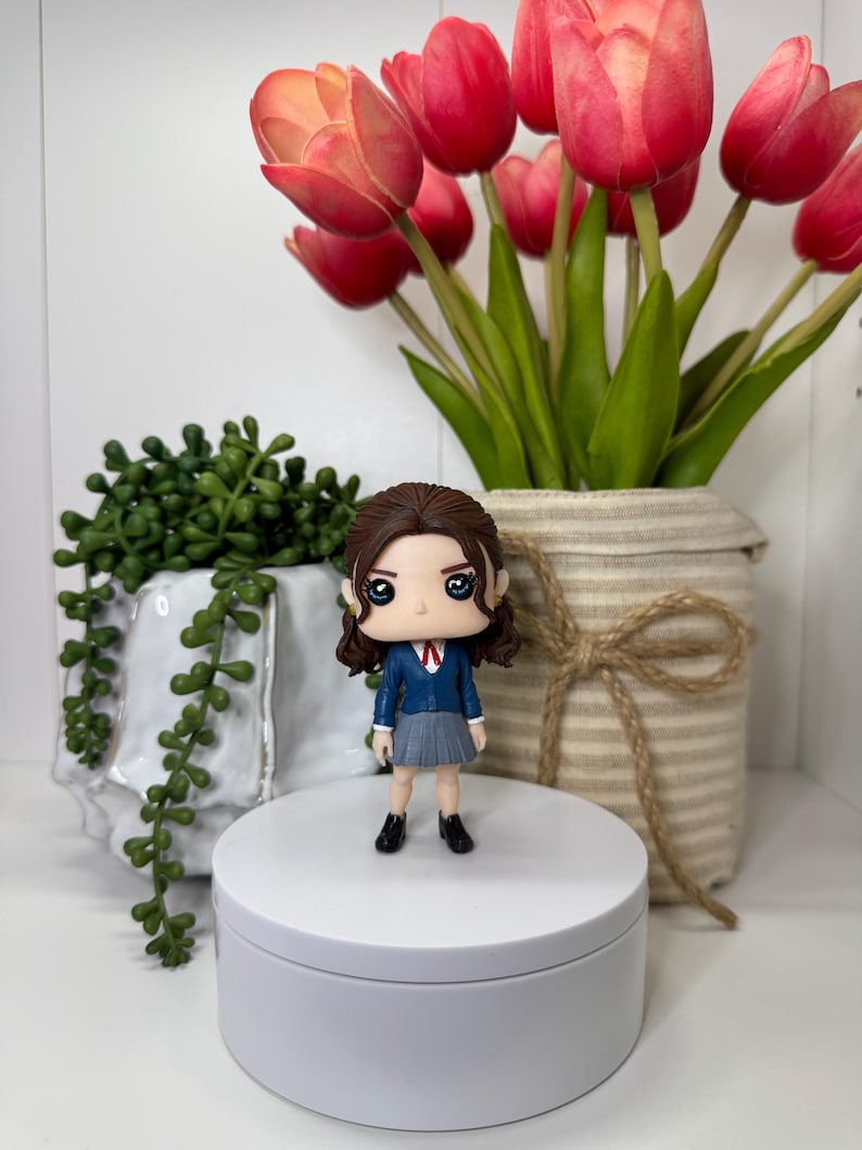 K&ouml;nnte beinhalten: Eine Figur einer Person in einem blauen Blazer, grauem Rock und schwarzen Schuhen steht auf einer wei&szlig;en Plattform. Die Figur hat braune Haare und gro&szlig;e Augen. Rote Tulpen in einem beigen Topf und gr&uuml;ne Pflanzen sind im Hintergrund.