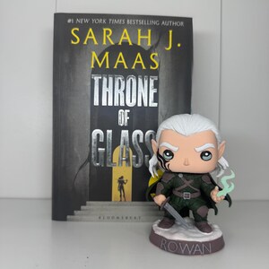 K&ouml;nnte beinhalten: Ein Buch mit dem Titel "Throne of Glass" von Sarah J. Maas steht neben einer Rowan Funko Pop Figur. Das Buch hat einen dunklen Einband mit silbernem Titel. Die Funko Pop Figur zeigt eine Figur mit wei&szlig;em Haar und einem Schwert.