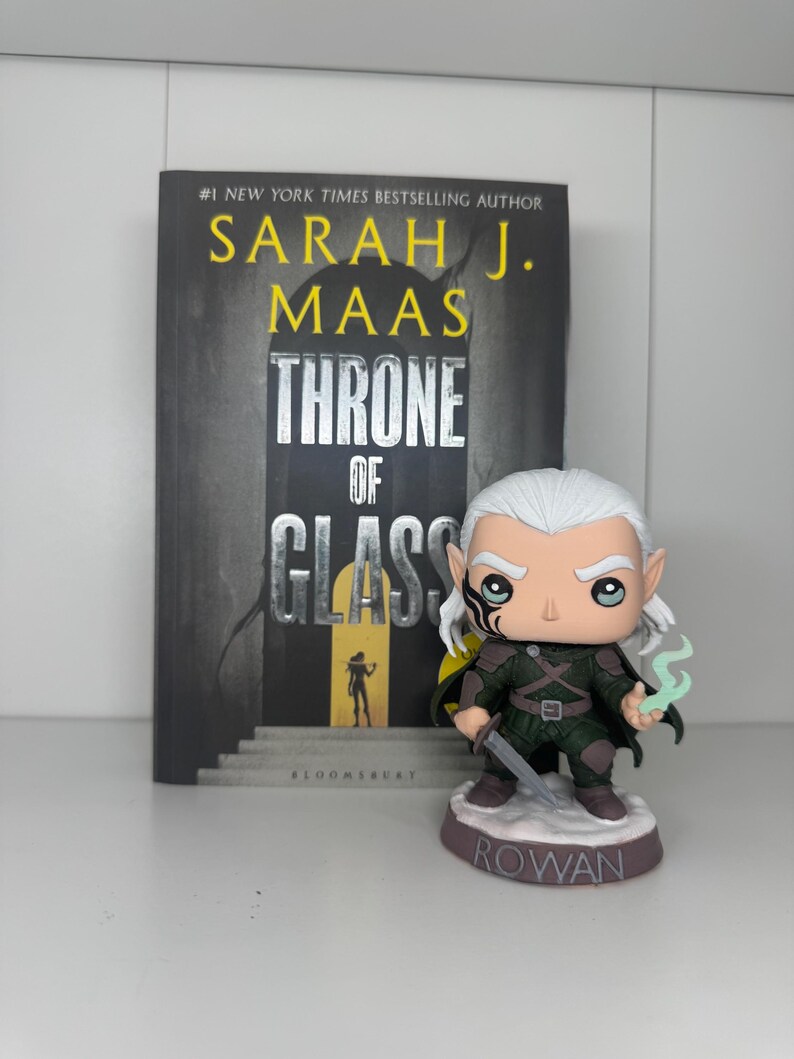 K&ouml;nnte beinhalten: Ein Buch mit dem Titel "Throne of Glass" von Sarah J. Maas steht neben einer Rowan Funko Pop Figur. Das Buch hat einen dunklen Einband mit silbernem Titel. Die Funko Pop Figur zeigt eine Figur mit wei&szlig;em Haar und einem Schwert.