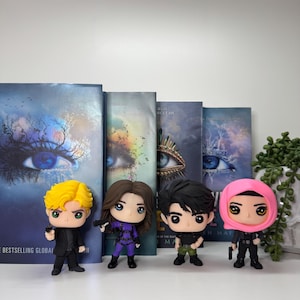 Pu&ograve; includere: Quattro personaggi Funko Pop di una serie di libri, in piedi davanti ai libri. Le figure includono un personaggio biondo in abito nero, un personaggio in tuta viola, un personaggio in camicia nera e pantaloni verdi e un personaggio in abito nero e velo rosa.