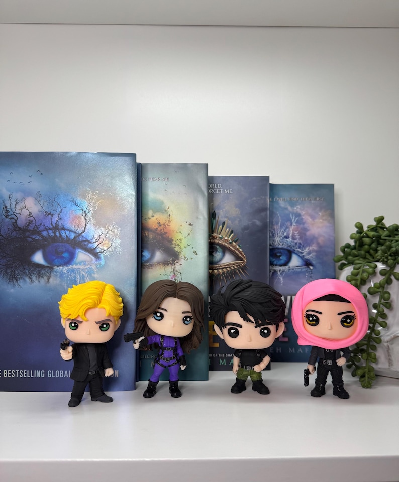 Pu&ograve; includere: Quattro personaggi Funko Pop di una serie di libri, in piedi davanti ai libri. Le figure includono un personaggio biondo in abito nero, un personaggio in tuta viola, un personaggio in camicia nera e pantaloni verdi e un personaggio in abito nero e velo rosa.