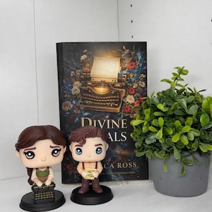 以下が含まれることがあります： 2体のFunko Popフィギュア、本、鉢植えの植物が置かれた棚。本の表紙には「Divine ALS」と書かれ、ヴィンテージのタイプライターのイラストが描かれています。フィギュアは男性と女性です。植物は灰色の鉢に入っています。