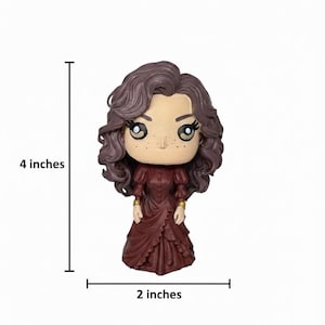 Może przedstawiać: Figurka kobiety z brązowymi kręconymi włosami i bordową sukienką. Figurka ma około 10 cm wysokości i 5 cm szerokości. Kobieta ma duże oczy i piegi. Złote akcenty na rękawach. Figurka na białym tle.